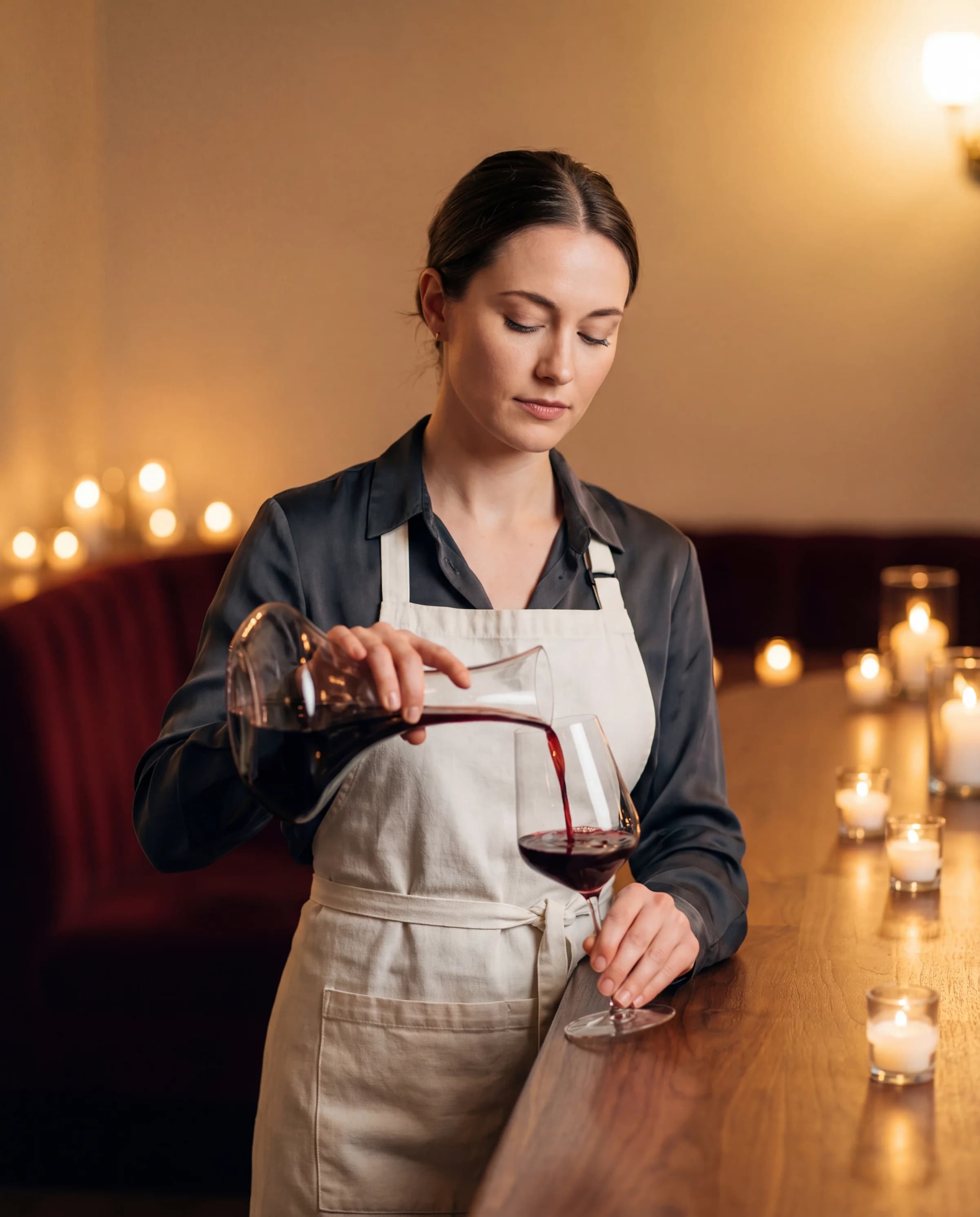 A poised sommelier in an ecru apron decanting Burgundy red wine into a Zalto glass on a candlelit walnut bar.